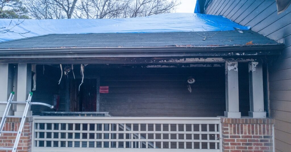 The outside of a home with the roof and porch damaged due to a recent fire. There’s a blue tarp over part of the roof.