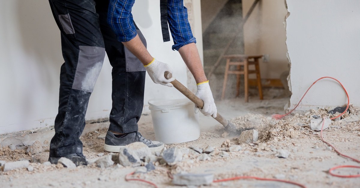 A worker wearing a flannel shirt and gloves using a shovel to clean up debris inside a building during a restoration project.
